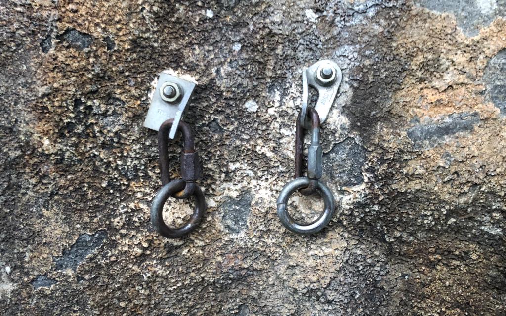 Two rings hanging from separate bolts. The bolt on the left is square and has 'Troll' stamped on it, the maillon and ring connected are rusty. The bolt on the right is rounded, the maillon is rusty, and the ring looks shiny.
